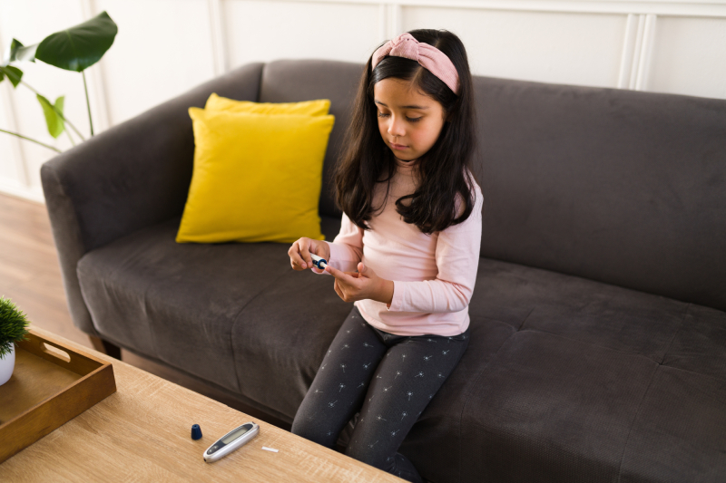 Image of a girl sitting at home in a couch, checking her glucose levels using blood testing strips and lancets.