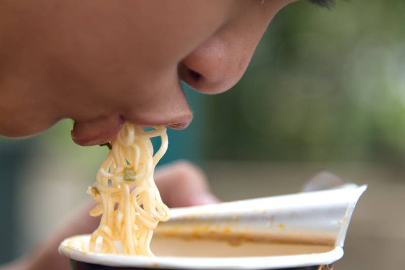 A boy is bending down and eating instant noodles with his mouth, without using a spoon.