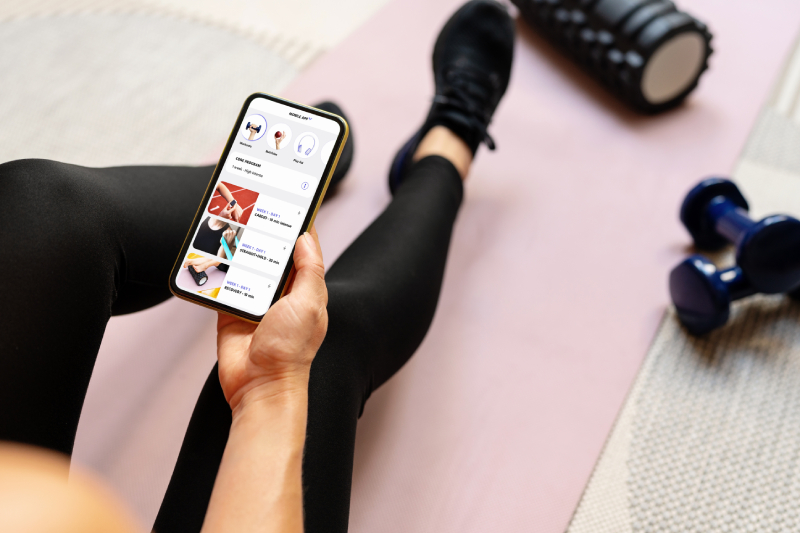 Top side view if a woman sitting on a yoga mat in a gym, holding a mobile displaying a fitness app and dumb bells placed near the mat.