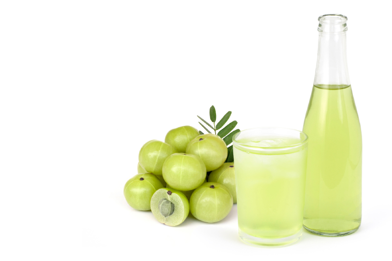 Few Indian gooseberries along with its leaves, a glass and bottle of gooseberry juice kept on a white background.