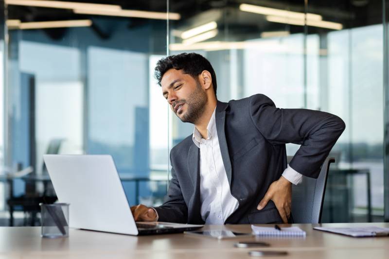 A businessman with pain sits at his office laptop, hands positioned behind his back.