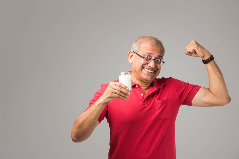 A senior man poses for a photo, holding a glass of juice and raising his arm happily.