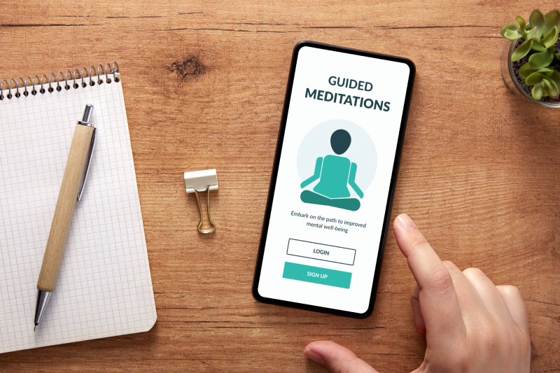 A pen rests on the notepad on the table, with a mobile phone displaying an open meditation app and a small indoor plant next to it.