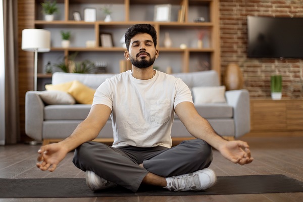 Image of a young man in exercise outfits doing meditation sitting on a mat inside a living room.