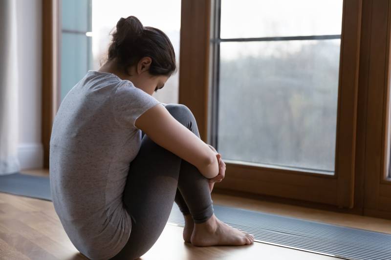 A female is seen from the back, sitting with her knees bent in front of a glass window, appearing to be in a bad mood.