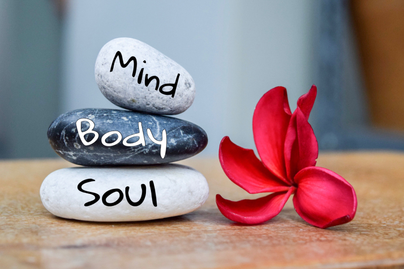 A brown table with white,black and grey stone arranged on top of the other with the words body,soul and mind written on it and a red plumeria flower kept beside it.