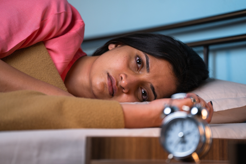 A young woman is sleeping on her bed, under a cover, with an alarm clock beside her on the table.