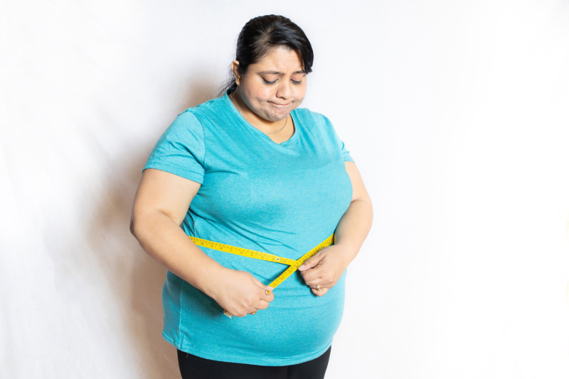 A sad overweight woman standing in front of a white background, checking her waist size with a measuring tape held around her waist.