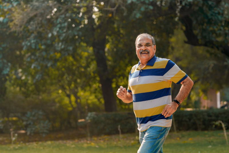 An elderly man with a smiling face jogging at a park with blurred images of trees at the background.