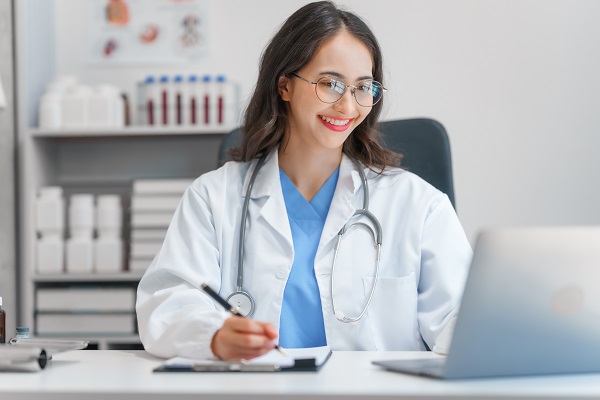 A female doctor sitting inside a consulting room with a smiling face doing an online consultation and writing down the prescription for the patient.