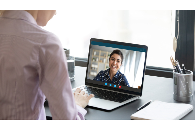 Rear view of a woman sitting in front of a laptop communicating with another woman over a video call and a glass of water, a pen holder and a note book is kept near it.