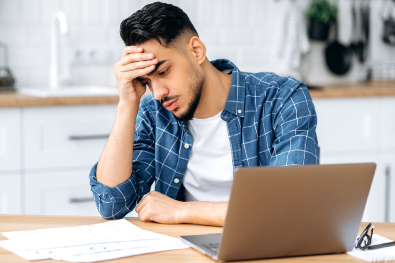 A stressed and frustrated young man looking at a graph kept before him along with a laptop,writing pad and spectacles.