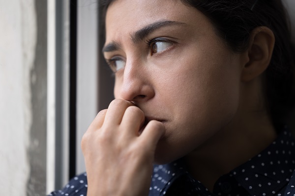 Close up view of a woman with a worried face standing near a window biting her nails, looking outside in some deep thought.