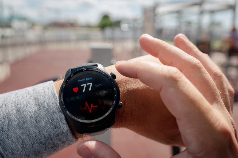 A persons hand touching his smart watch worn on the other hand, displaying a health tracker showing the heart rate on the screen and blurred inside images of an athletic stadium at the background.