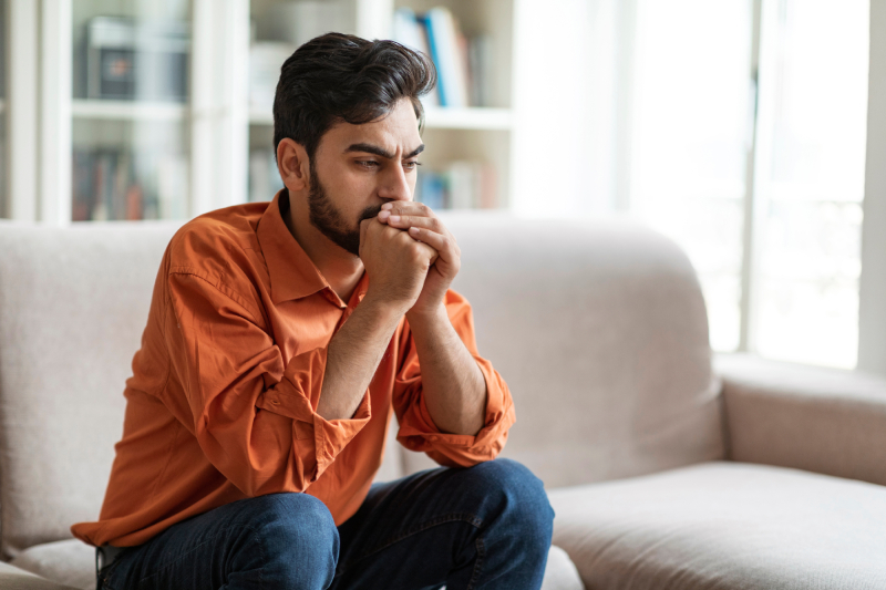 A tense young man sits on a sofa at home, leaning on his hands as he thinks.