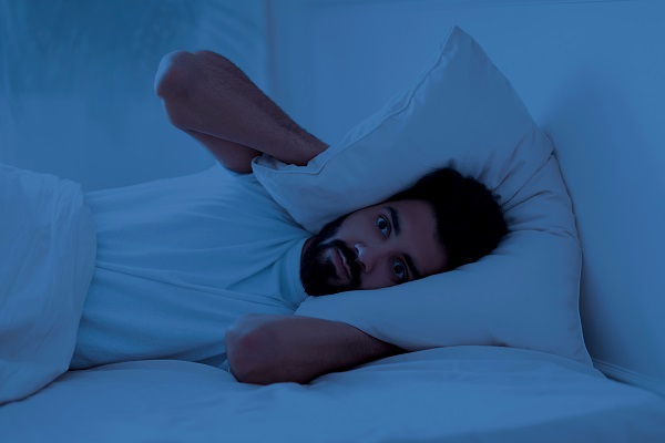 A young man, feeling anxious, is awake at night with a pillow over his head.