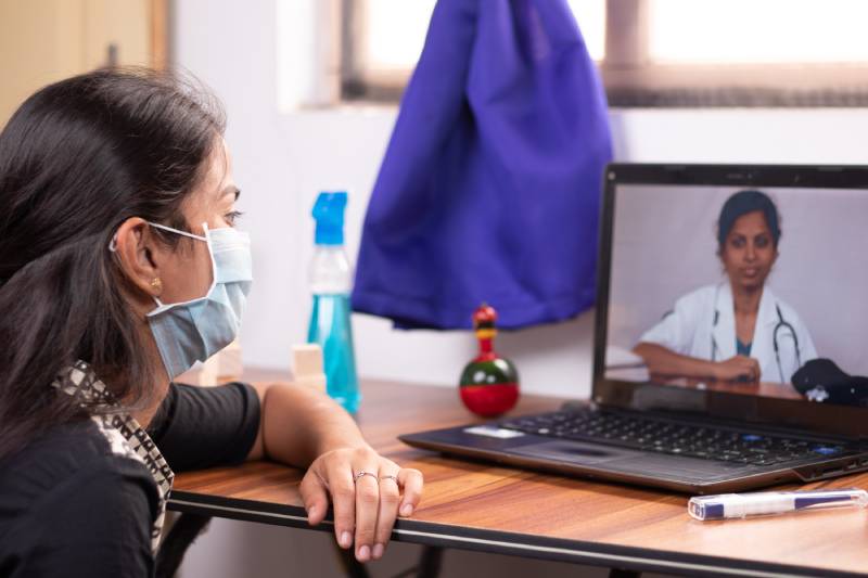 A young girl in a mask consults a doctor through video conferencing on her laptop, with sanitizer and a thermometer nearby on the table.