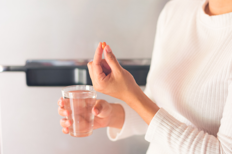 A woman holds an oval-shaped orange tablet in her left hand and a glass of fresh water in the other.