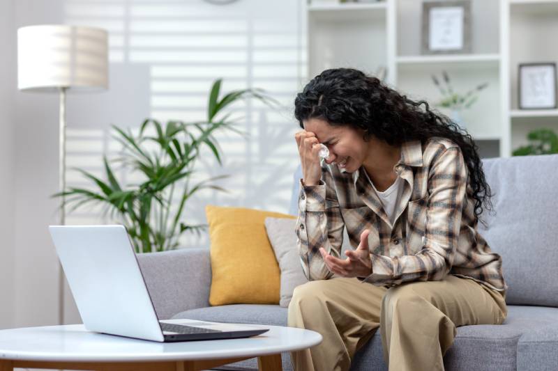 A woman cries sitting on a lounge sofa during an online consultation through a laptop before her.