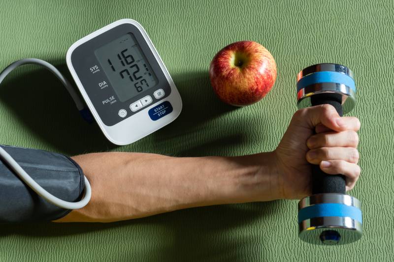 A hand with a BP monitor cuff , holding a dumb bell and an apple kept near it.