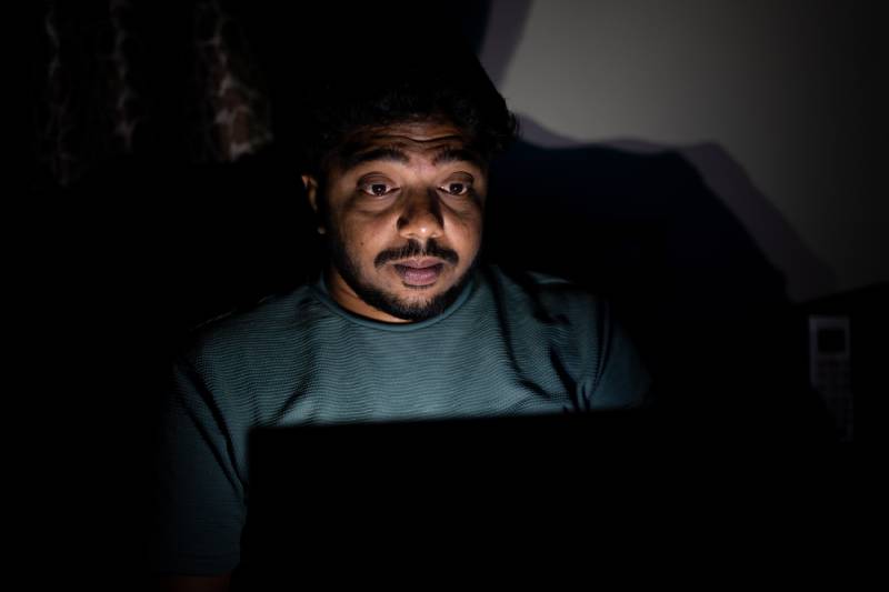 An overstressed man looking at his laptop screen sitting in a dark environment.