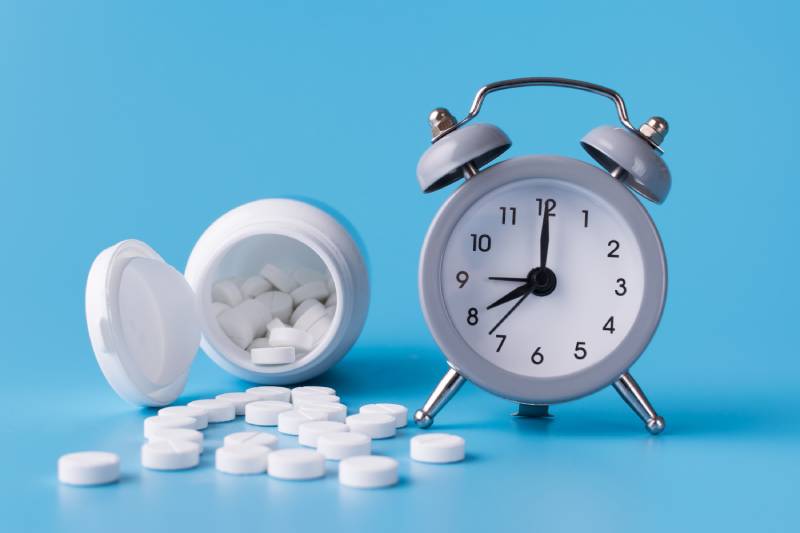 An alarm kept near an open bottle of tablets kept on a blue background.