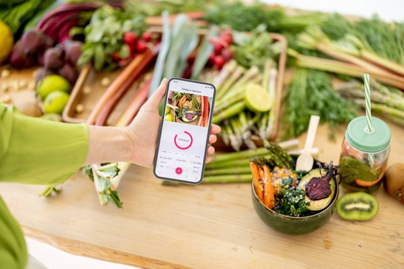 A female holding a mobile displaying an app, tracks the nutritious value of food items kept on a table.