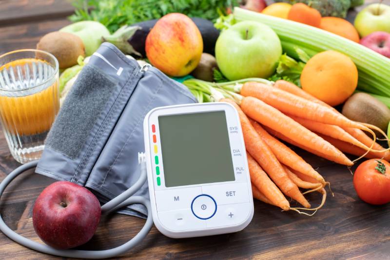 Digital BP monitor kept on a heap of fresh fruits and vegetables arranged on a wooden table.