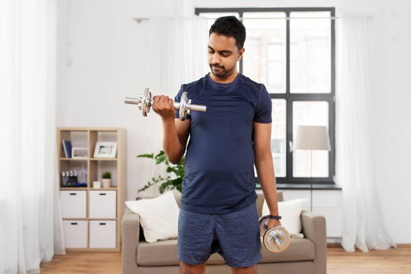 A man exercising with dumb bells standing inside a living room.