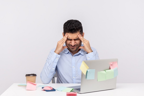 A stressed male employee holds his head, suffering from a headache due to workplace stress and burnout.