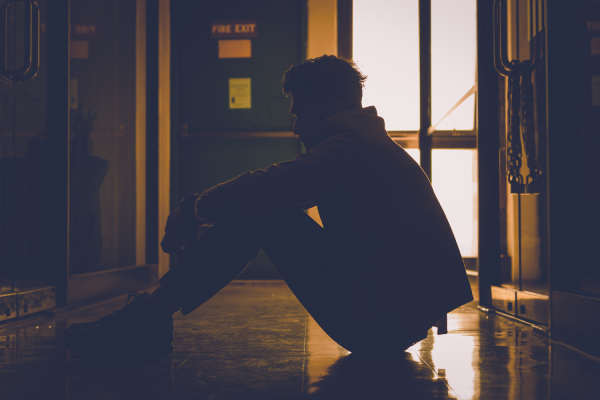 A depressed young man sits on the floor in darkness, reflecting the stigma and blame society places on mental health struggles.