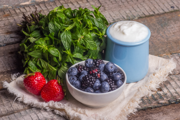 Fresh strawberries, blueberries, and blackberries in a bowl with mint leaves and a jar of yogurt on a rustic wooden table.