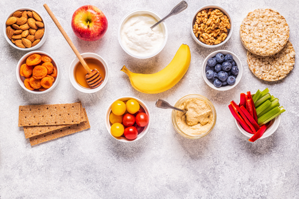 Healthy snacks like nuts, fruits, yogurt, and veggies on a gray background, provide essential fats for growth and vitamin absorption.