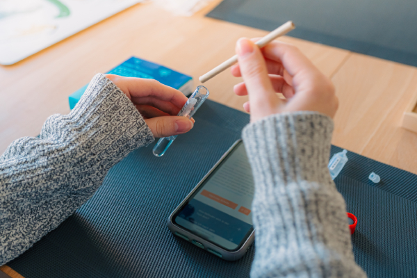 A person in a gray sweater collects a DNA sample using a saliva testing kit, with a smartphone showing instructions on the table.