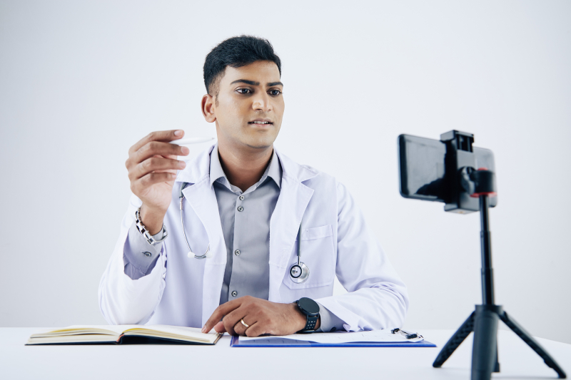 Doctor in a white coat performing a remote consultation using software, seated at a desk with a smartphone tripod.