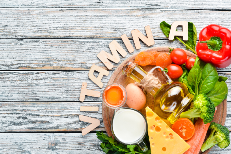 Vitamin A-rich foods like carrots, spinach, eggs, and tomatoes on a wooden surface, with 'VITAMIN A' spelled out in wooden letters.