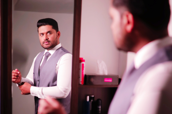 Man in grey vest and tie adjusting his attire in the mirror, with a digital clock on the wall, reflecting confidence.