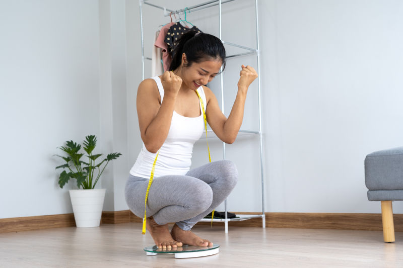 A happy woman with a measuring tape, sitting on a scale, celebrating the results of her slimming journey.